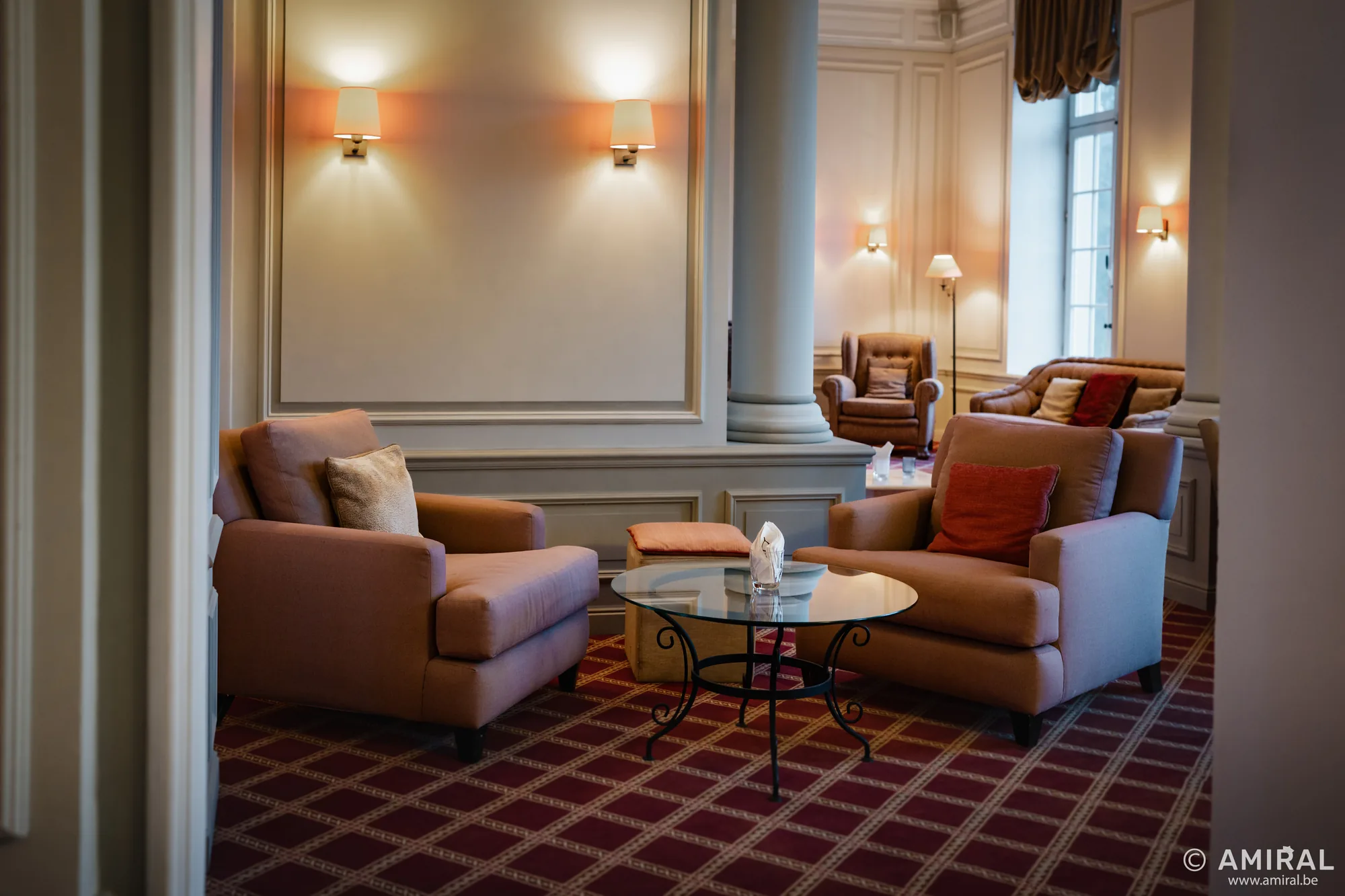 Salon élégant avec canapés beiges et fauteuils orange autour d'une table basse, moquette bordeaux à motifs.