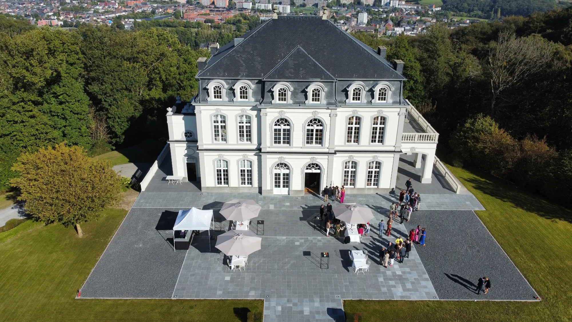 Grande demeure blanche à mansardes vue du ciel, entourée de pelouse verdoyante et de véhicules stationnés