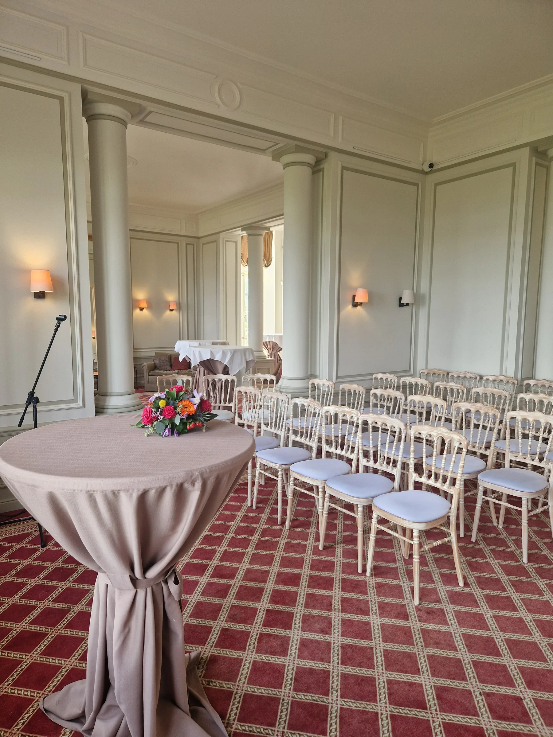 Salle de réception avec chaises blanches alignées, moquette rouge à motifs et table cocktail drapée
