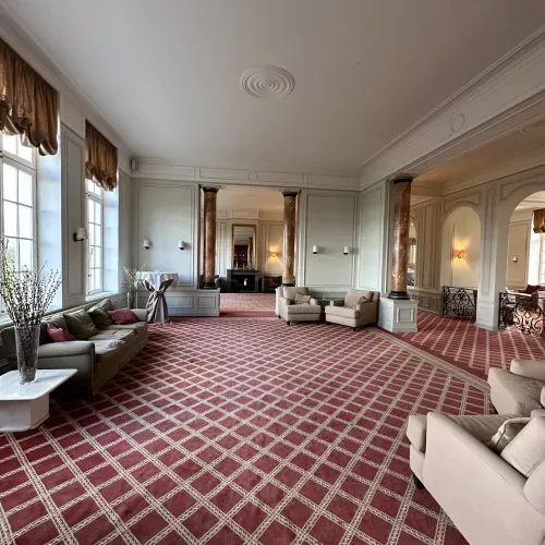 Grand salon avec moquette à motifs écossais bordeaux, canapés beiges et grandes fenêtres lumineuses