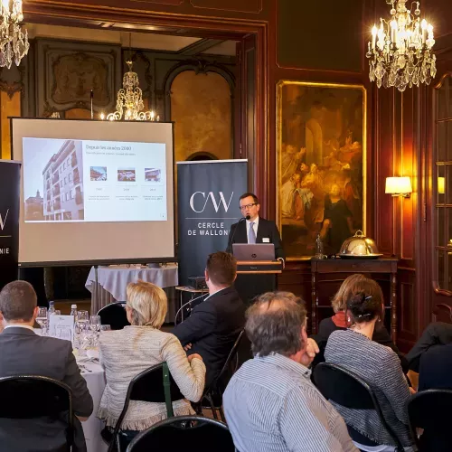 Conférence dans une salle élégante avec lustre, public assis écoutant un présentateur devant un écran.
