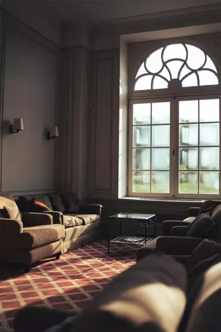 Salon lumineux avec canapé beige, fenêtre cintrée et lumière naturelle créant des ombres sur le sol.